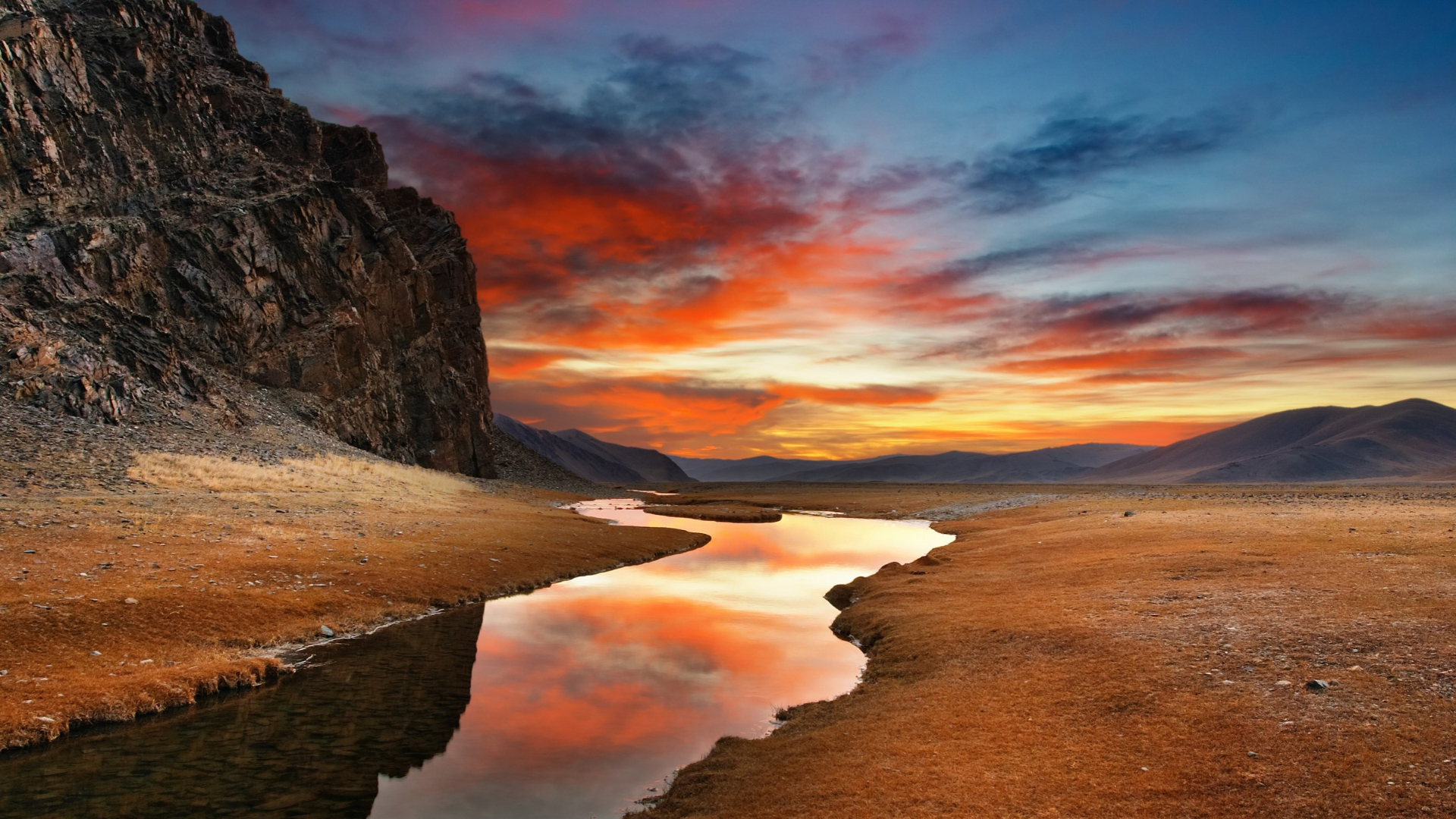 A dramatic sunset over a vast Mongolian steppe, with a winding river reflecting vivid orange and red clouds beneath rugged cliffs and distant mountains.