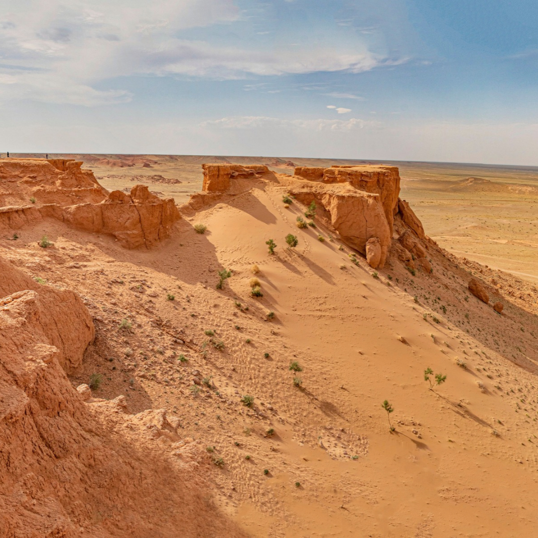 Red sandstone cliffs and eroded rock formations rise above the vast, arid landscape of Mongolia’s Gobi Desert beneath a pale blue sky.