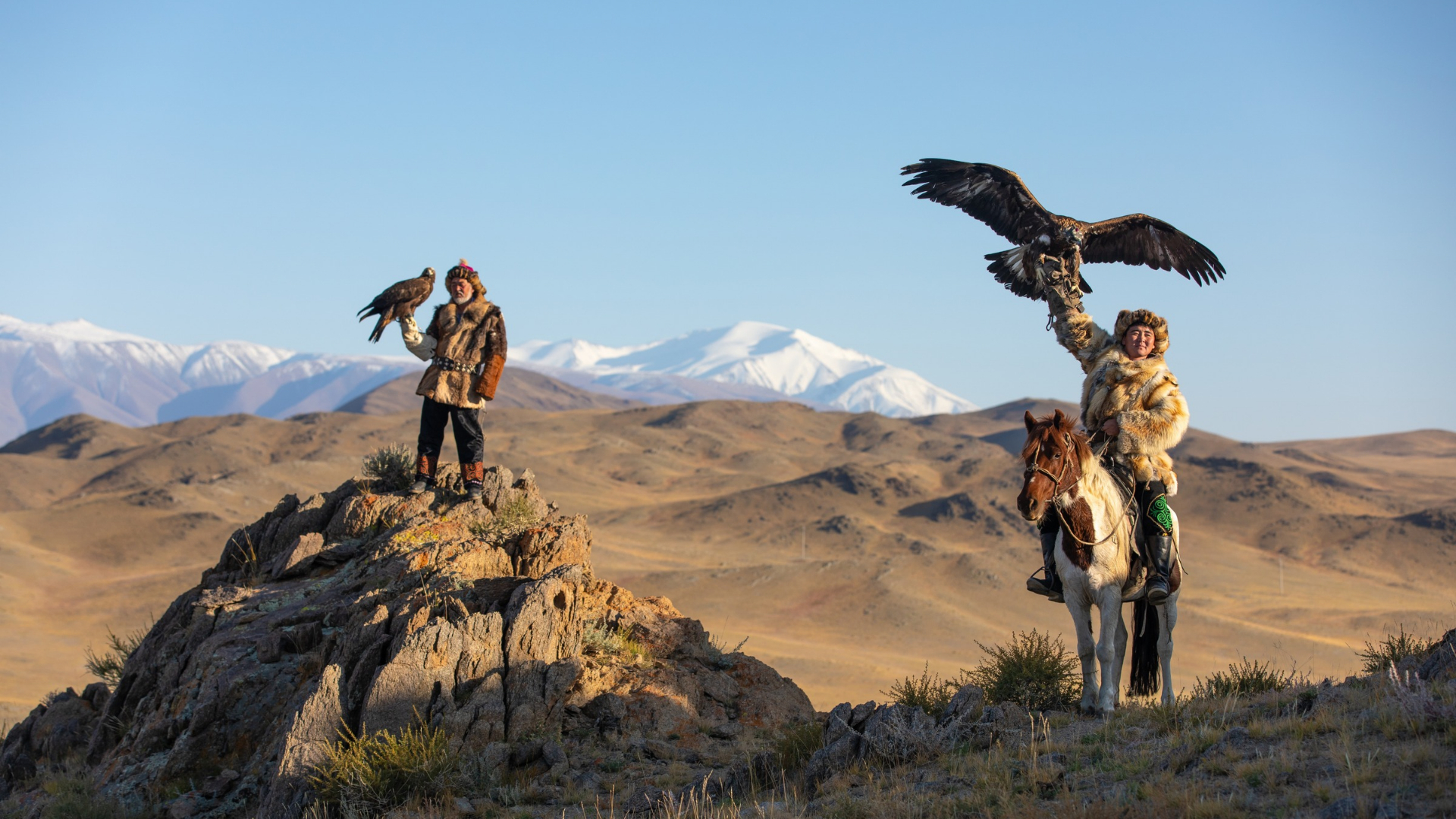 Two traditional Mongolian eagle hunters pose with golden eagles in a rugged mountain landscape, one standing on a rocky outcrop and the other seated on horseback.