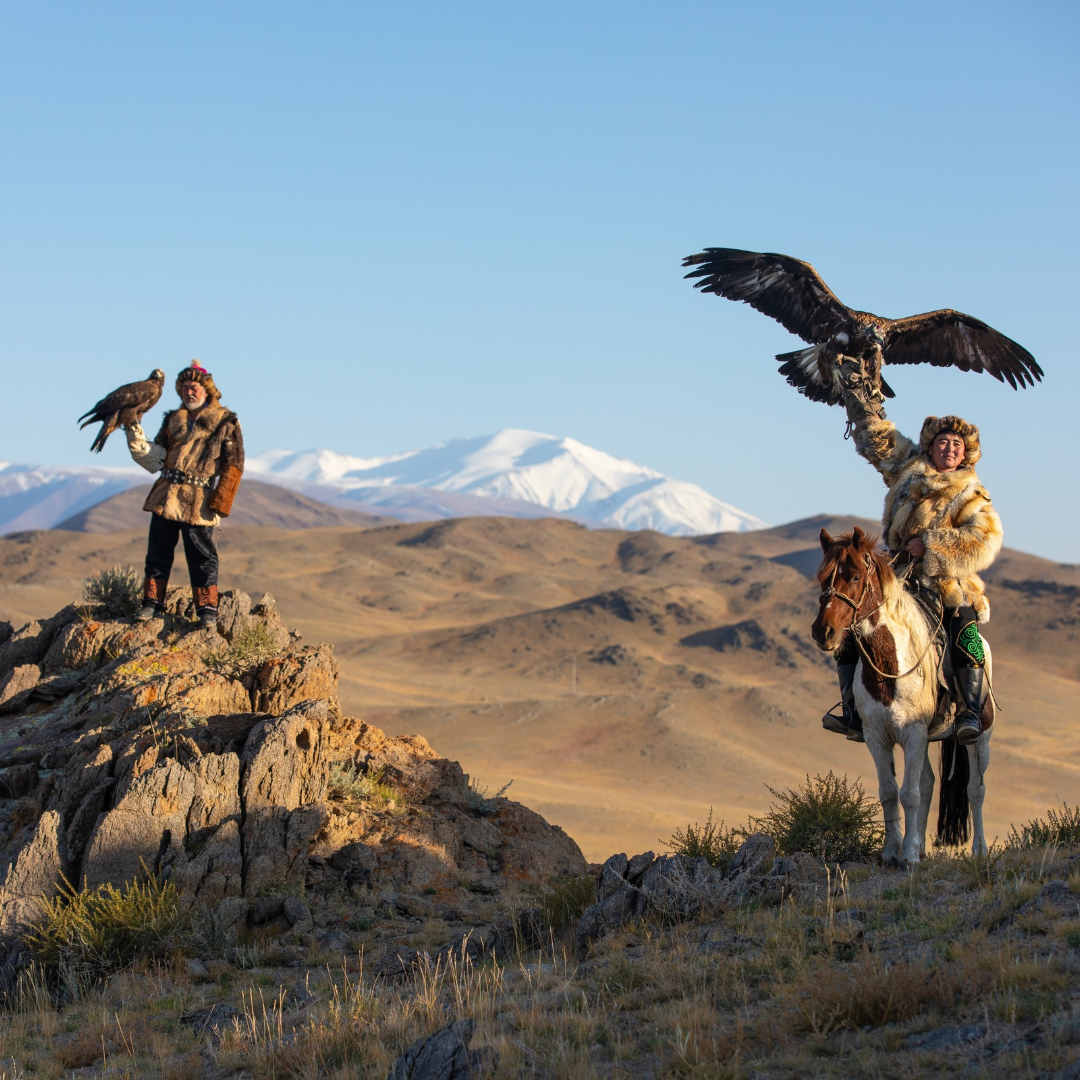Two traditional Mongolian eagle hunters pose with golden eagles in a rugged mountain landscape, one standing on a rocky outcrop and the other seated on horseback.