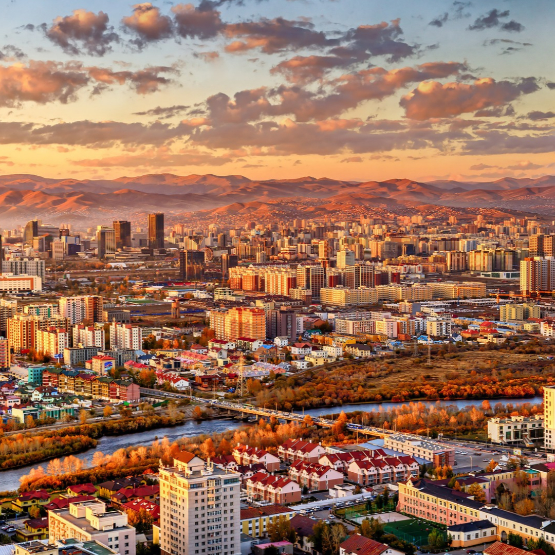 Panoramic view of Ulaanbaatar, Mongolia, with dense city buildings along a river, surrounded by rolling mountains glowing in warm sunset light.