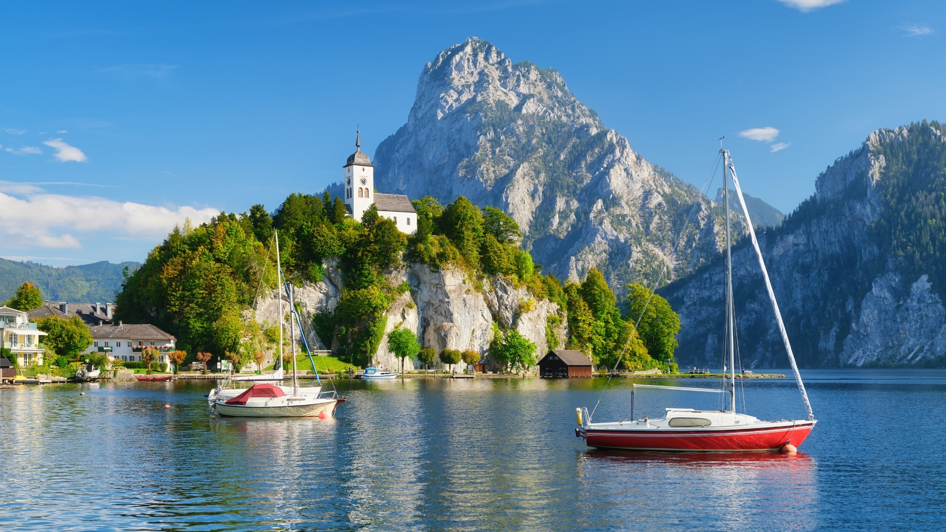 The image depicts a serene lake scene in Switzerland, featuring two boats in the foreground, a small church on a rocky outcrop surrounded by lush greenery, and majestic mountains in the background under a clear blue sky.