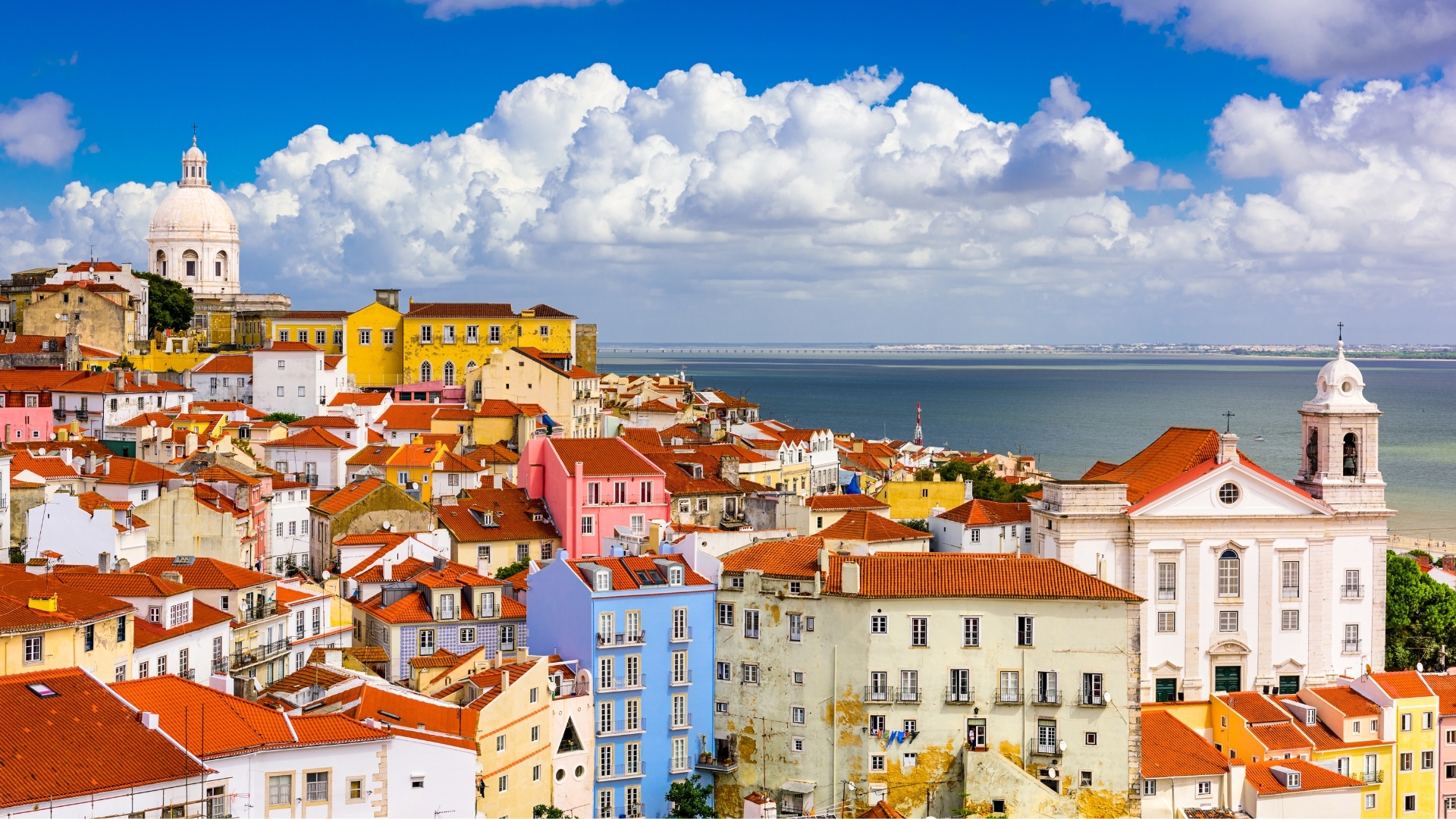 The image depicts a vibrant cityscape of Alfama, Lisbon, showcasing colorful buildings with red rooftops, a prominent dome structure, and a view of the river under a bright blue sky with fluffy clouds.