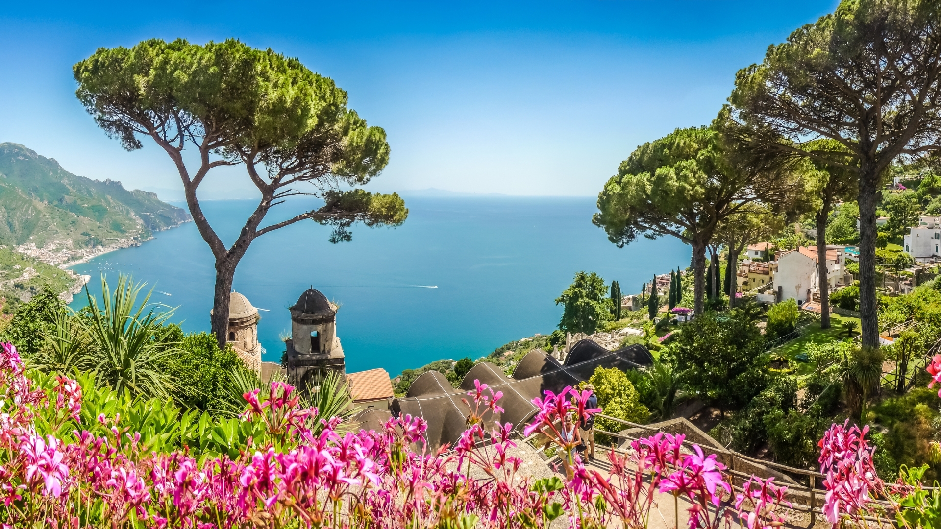 The image depicts a stunning view of the Amalfi Coast in Campania, Italy, showcasing vibrant pink flowers in the foreground, lush greenery, and a serene blue sea under a clear sky. The coastline features charming houses and steep cliffs, with tall trees adding to the picturesque landscape.