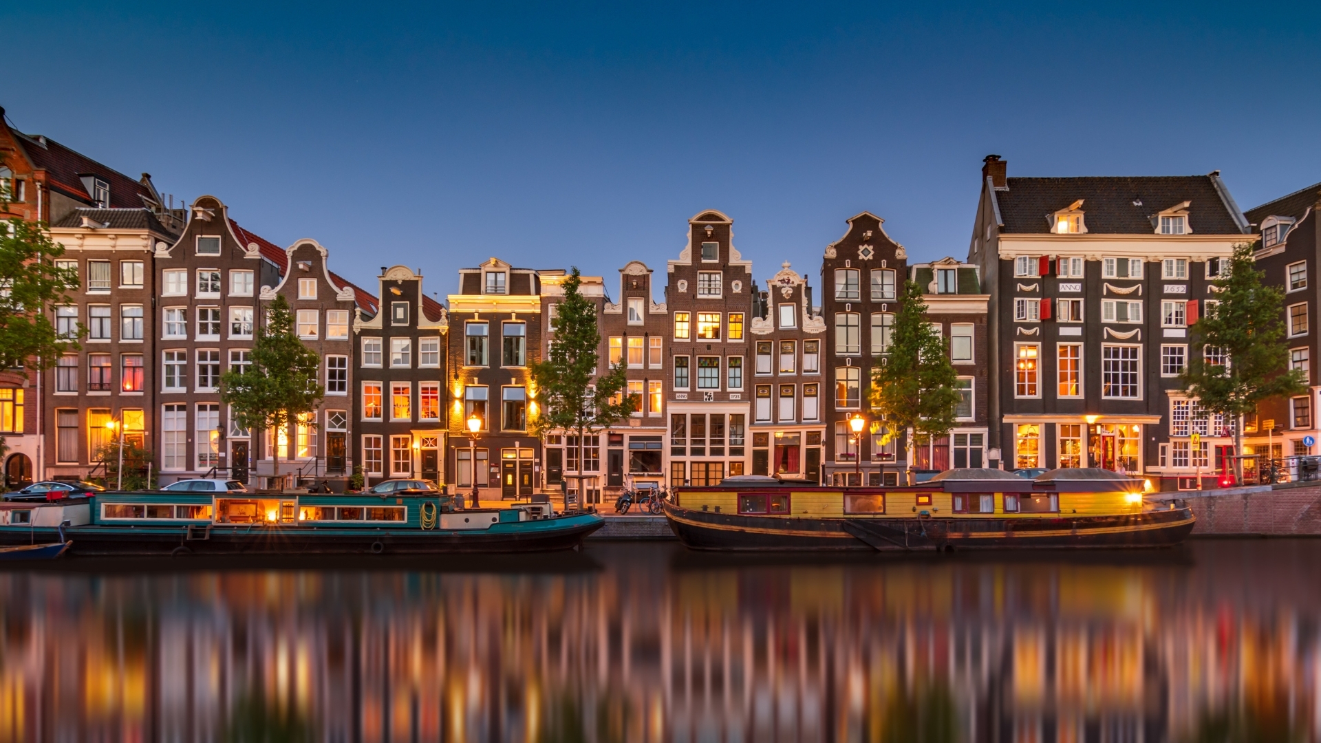 The image depicts a scenic view of Amsterdam's canals, showcasing traditional Dutch architecture with tall, narrow houses lining the water. The buildings feature gabled roofs and large windows, reflecting beautifully in the calm water of the canal. The scene is illuminated by warm lights, creating a charming evening atmosphere.