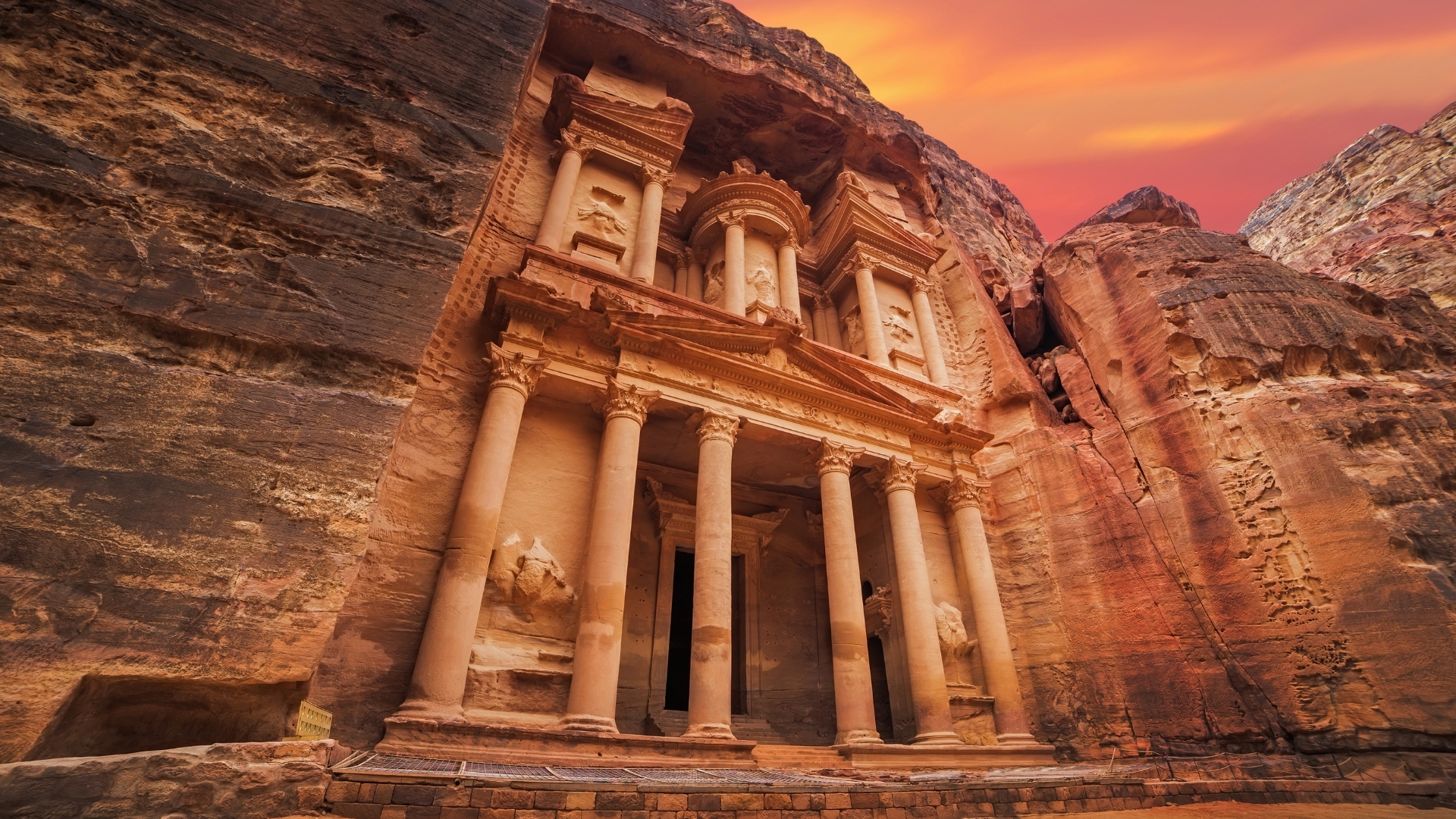 The image depicts the ancient temple known as Al-Khazneh, located in Petra, Jordan. It features grand columns and intricate carvings, set against a backdrop of towering rock formations and a vibrant sunset sky.