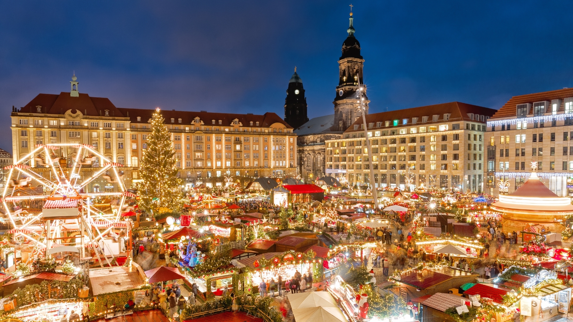 The image depicts a vibrant Christmas market in Dresden, showcasing festive decorations, a large Christmas tree, and numerous stalls filled with holiday goods. The scene is illuminated with lights, creating a warm and inviting atmosphere, typical of traditional European winter holiday celebrations.