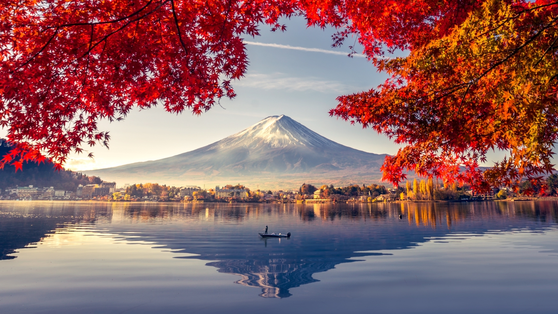 The image depicts a serene view of Mount Fuji surrounded by vibrant autumn foliage, with red leaves framing the scene. A calm lake reflects the mountain and the colorful trees, while a small boat with a person is seen on the water, creating a peaceful atmosphere.