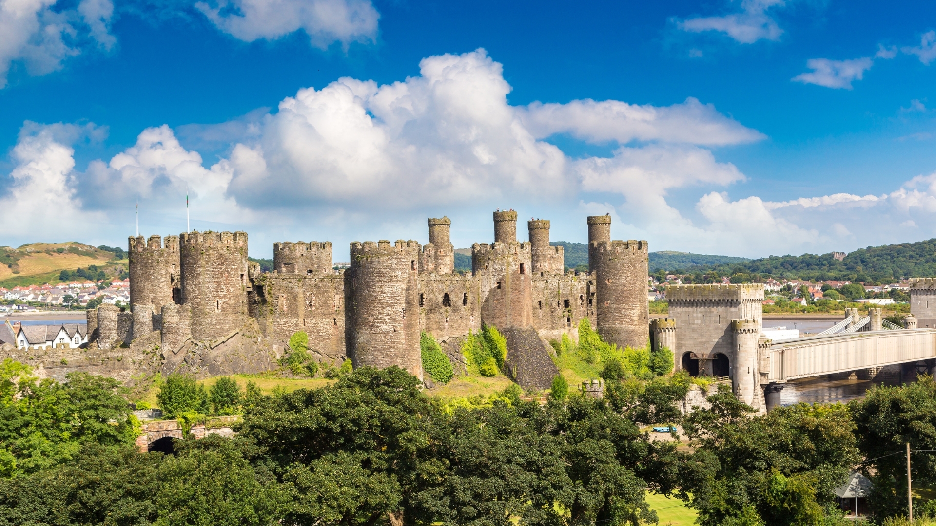 The image depicts Conwy Castle in Wales, showcasing its impressive stone walls and towers against a backdrop of blue skies and fluffy clouds. The castle is surrounded by lush greenery and is part of a scenic landscape that includes a bridge and distant hills.