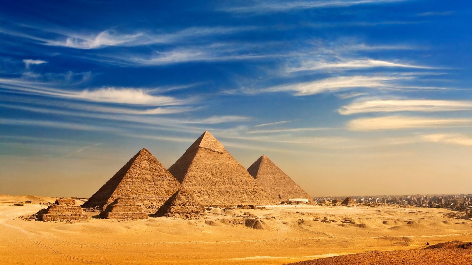 The image shows a panoramic view of the Giza Pyramids against a clear blue sky, with the cityscape of Cairo visible in the background. The pyramids, known for their ancient significance, are set in a desert landscape, highlighting their grandeur and historical importance.