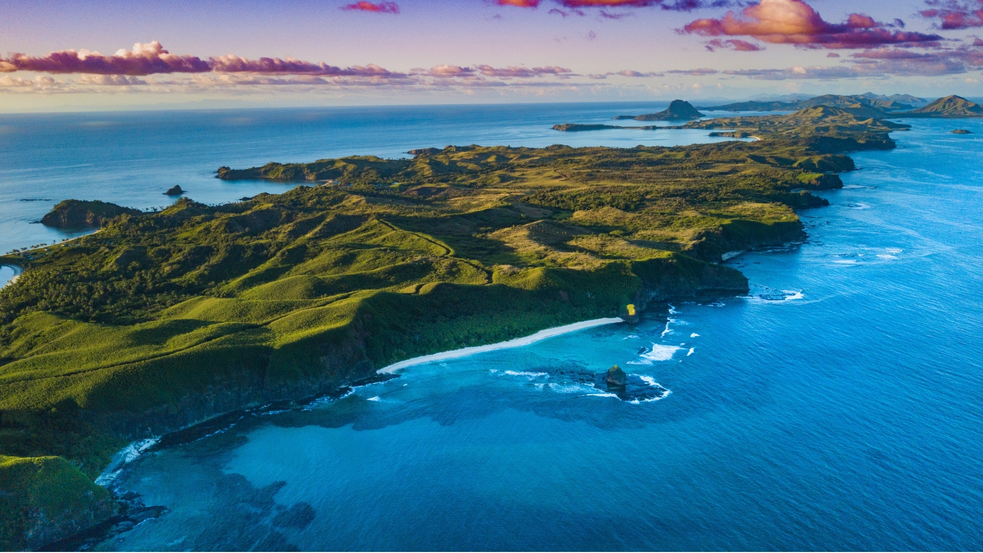 The image shows a stunning aerial view of a lush green island landscape surrounded by clear blue waters. The coastline features a sandy beach and rocky formations, with hills and mountains visible in the background under a colorful sky.