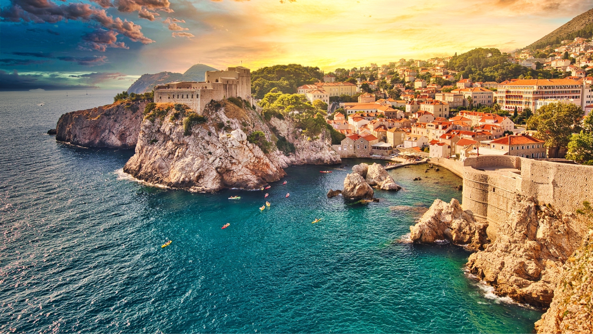 The image depicts a scenic view of Fort Lovrijenac, a historic fortress located on a rocky cliff overlooking the Adriatic Sea, with the city of Dubrovnik in the background. The scene is bathed in warm sunset colors, highlighting the vibrant rooftops of the city and the clear blue waters, while kayakers can be seen paddling near the shore.