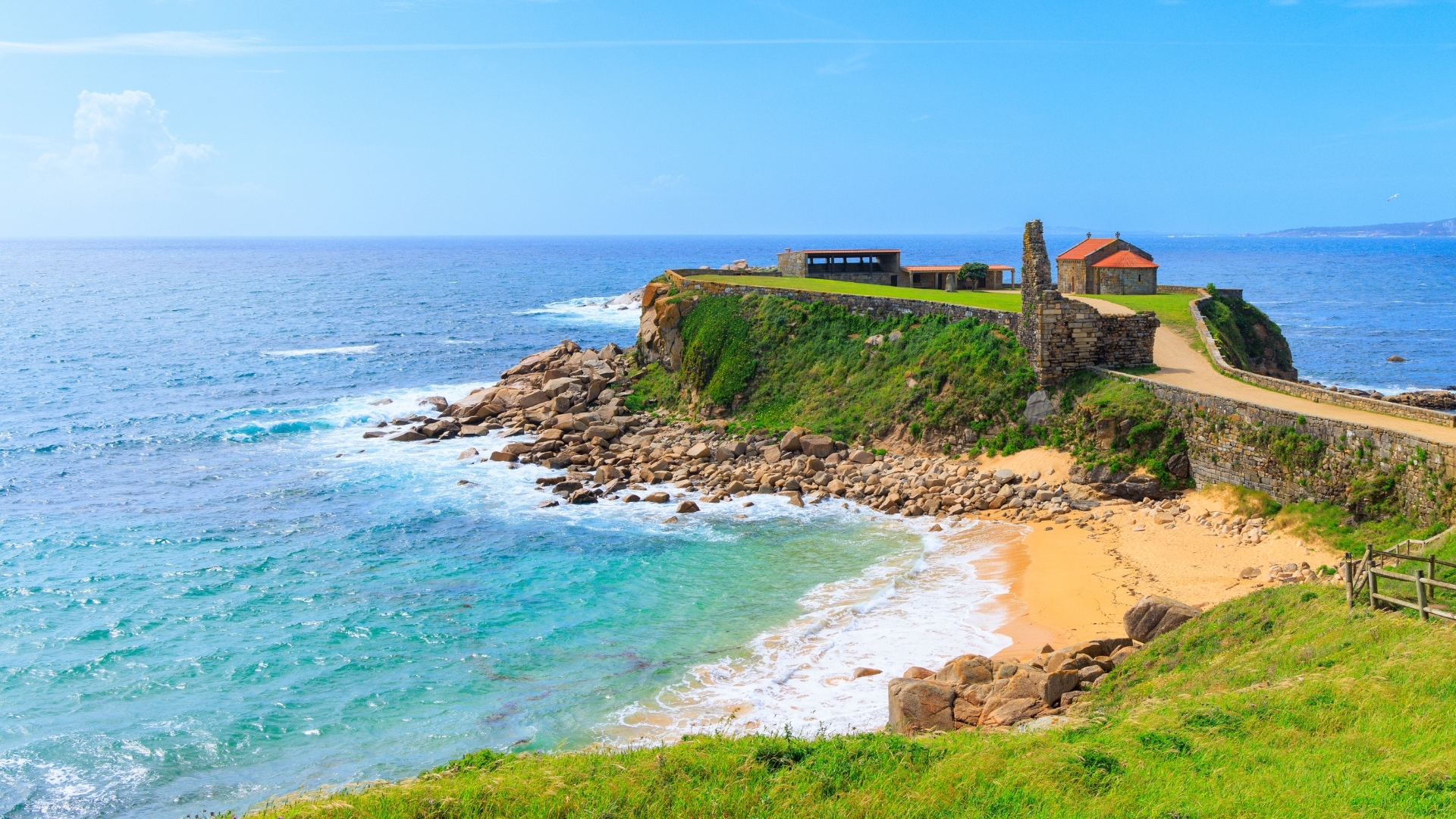 The image depicts a scenic view of the Atlantic Ocean from a coastal area in Galicia, Spain. It features rocky shores, a sandy beach, and a small stone building with a red roof, surrounded by lush greenery and a clear blue sky.