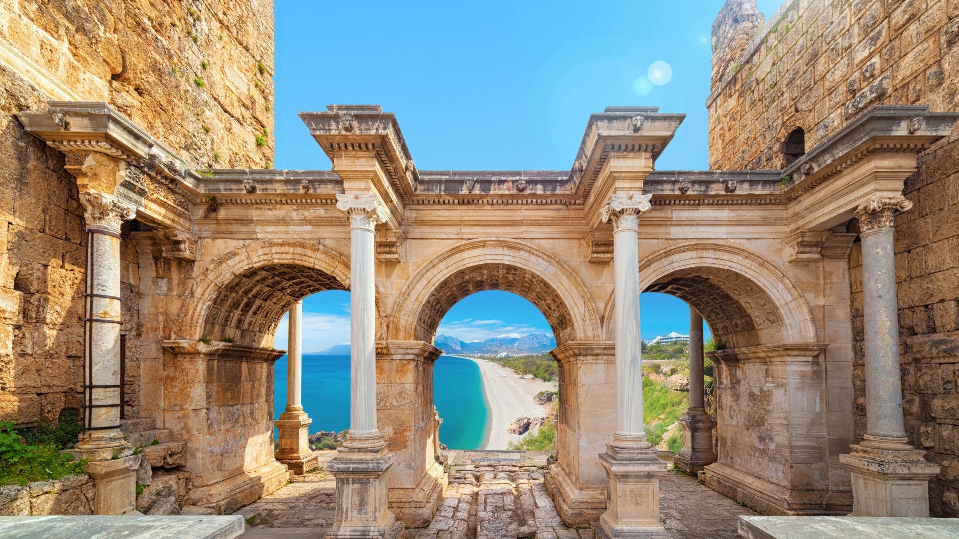 The image depicts Hadrian's Gate, an ancient Roman structure located in Antalya, Turkey. It features three grand arches supported by tall columns, with a view of a beautiful beach and turquoise sea in the background, showcasing a blend of historical architecture and natural beauty.
