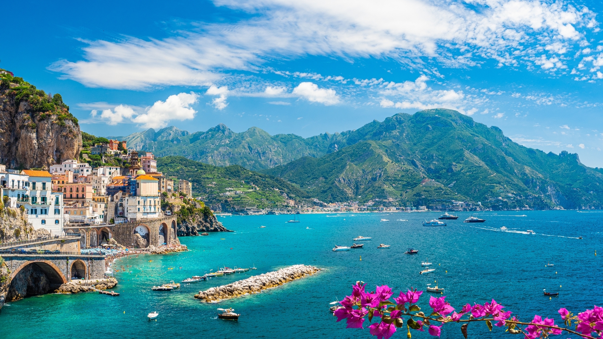 The image depicts a vibrant coastal landscape featuring the town of Atrani along the Amalfi Coast in Italy. The scene showcases colorful buildings perched on cliffs, surrounded by lush green mountains and a sparkling blue sea dotted with boats. Bougainvillea flowers in the foreground add a pop of color to the picturesque view.
