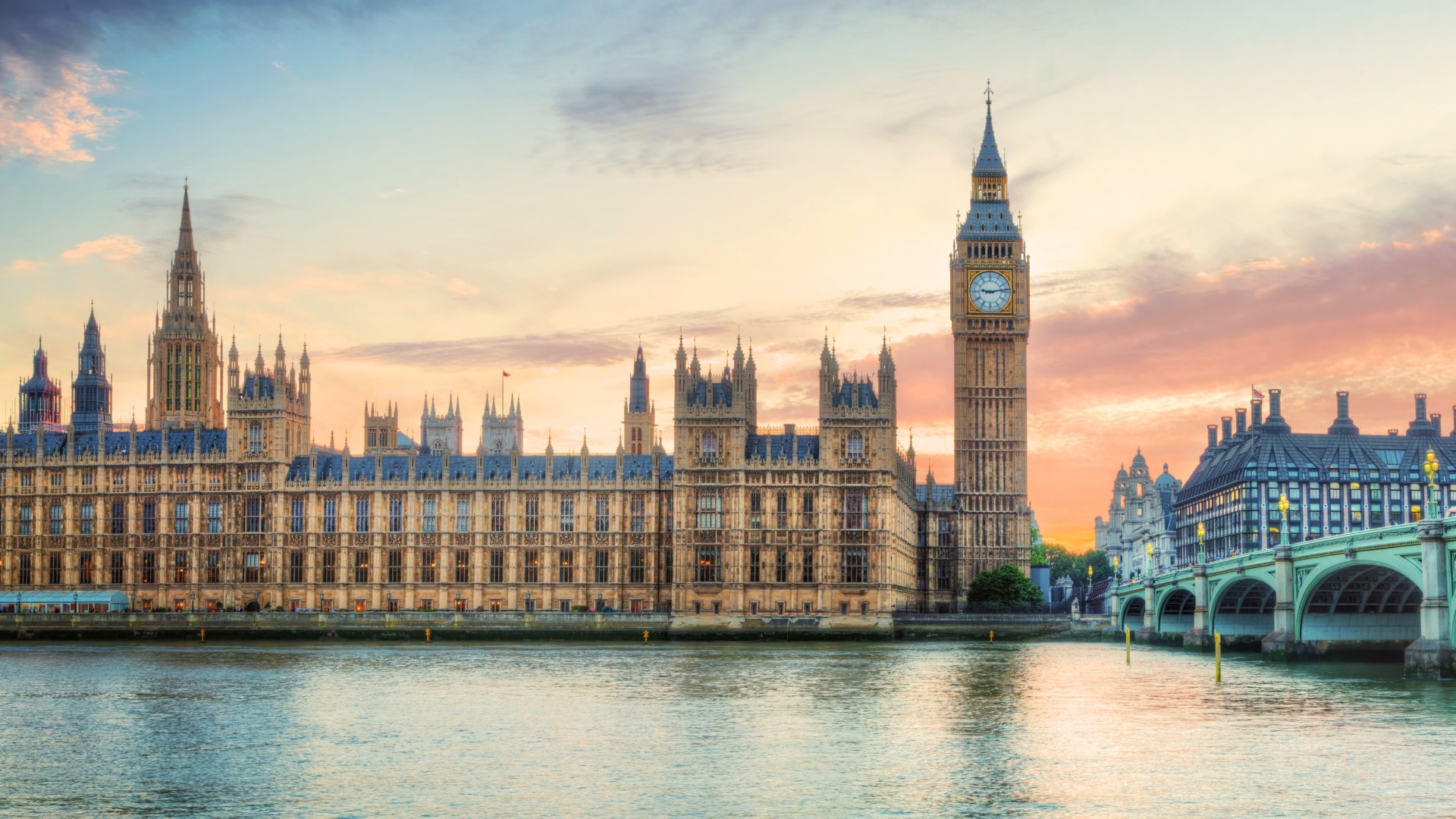 The image depicts a panoramic view of London at sunset, featuring the iconic Big Ben clock tower and the Palace of Westminster along the River Thames. The scene is illuminated by warm sunset colors, highlighting the architectural details of the buildings and the calm water of the river.