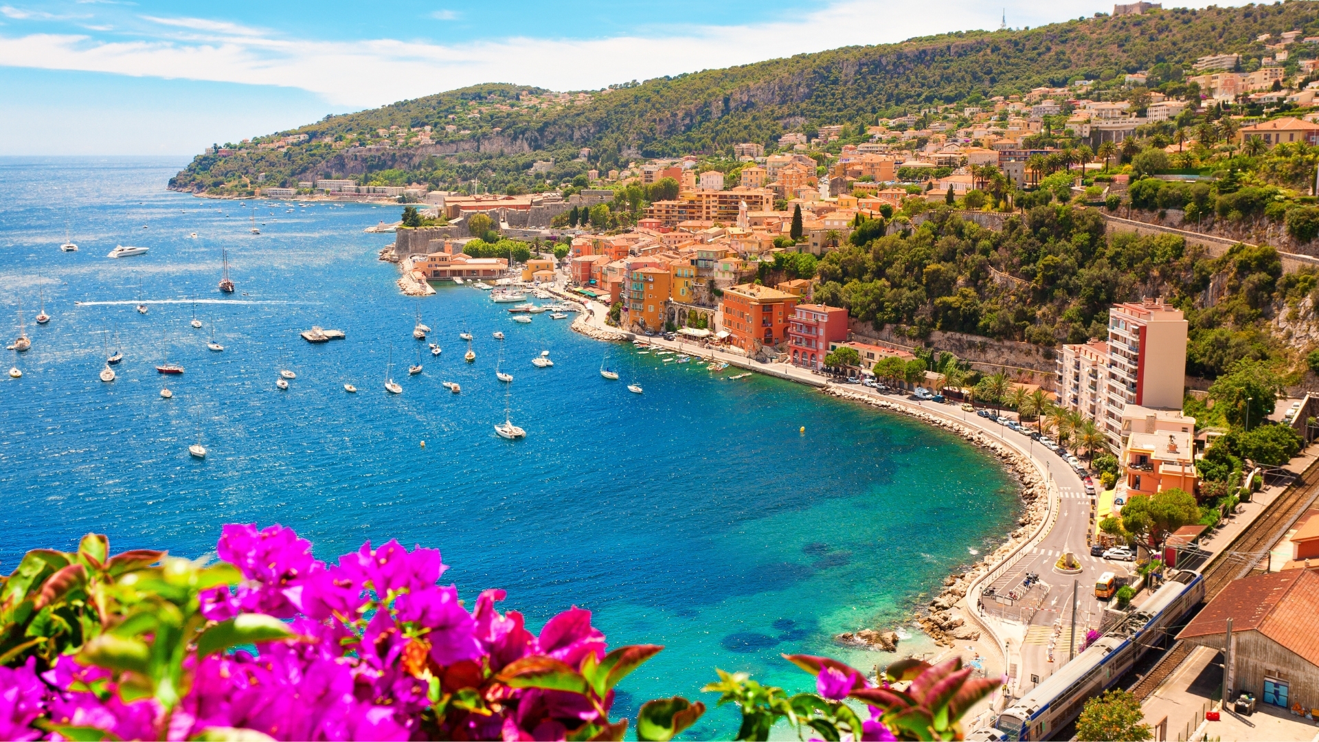 The image depicts a picturesque coastal view of Villefranche-sur-Mer, located on the French Riviera. It features a vibrant blue sea dotted with boats, colorful buildings lining the shore, and lush green hills in the background, creating a serene and inviting atmosphere.