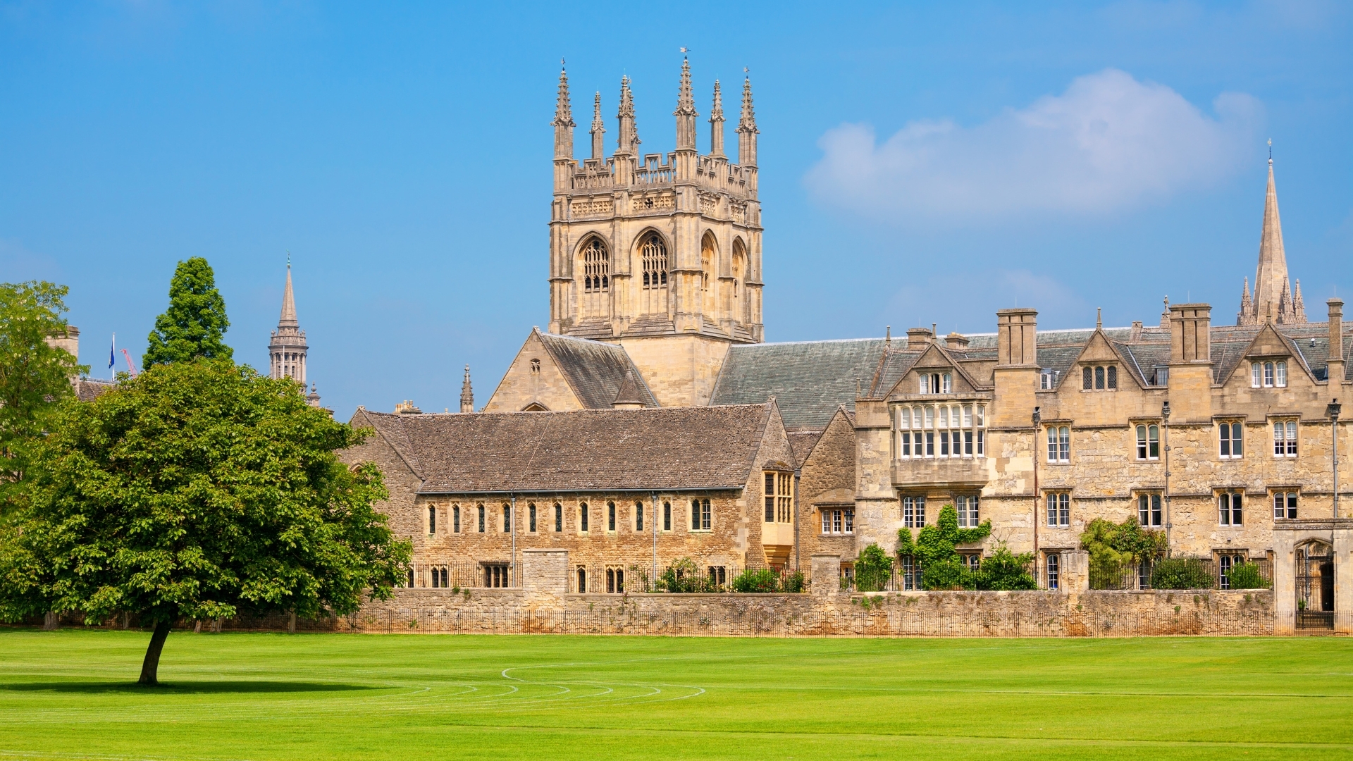 The image depicts Merton College, one of the colleges of the University of Oxford, UK. It features historic stone buildings with a prominent tower and spires against a clear blue sky, surrounded by a lush green lawn and trees.