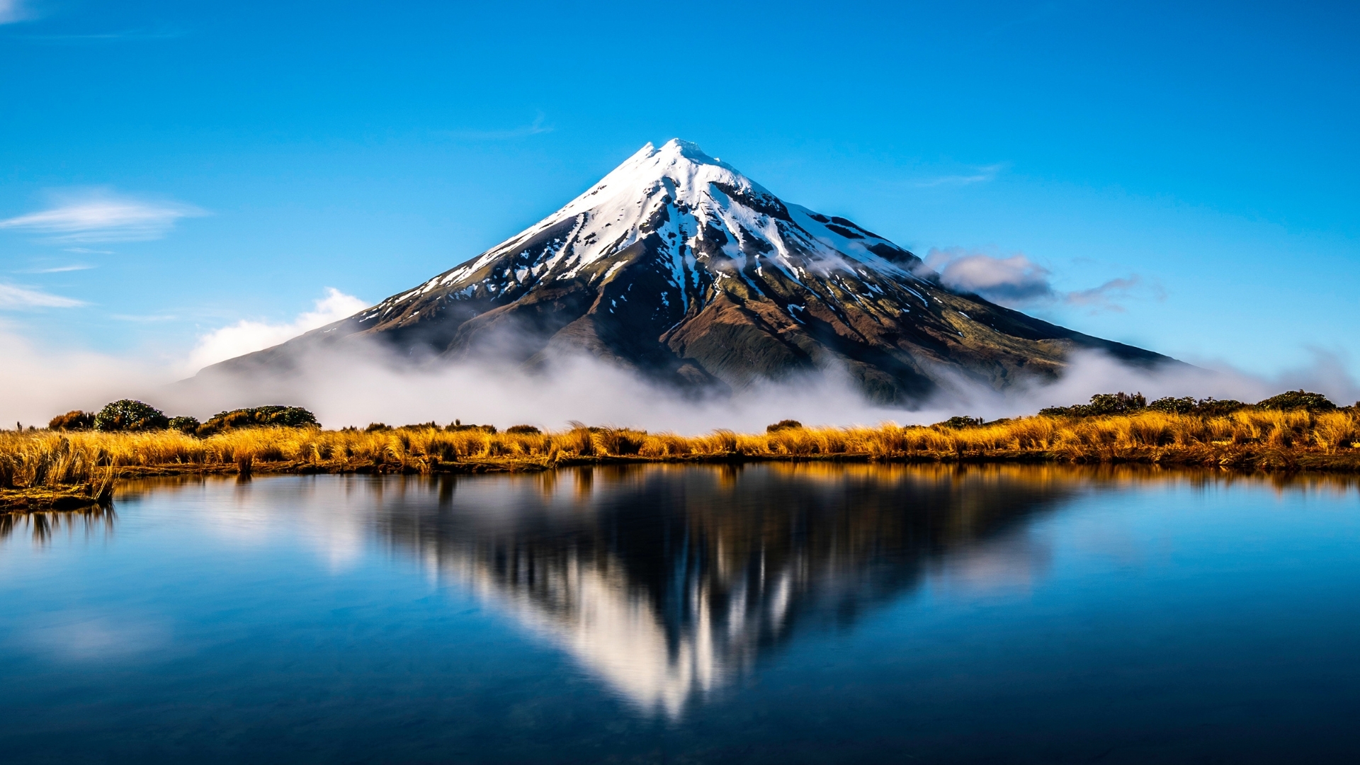 The image depicts Mount Taranaki in New Zealand, a snow-capped volcano reflected in a serene lake surrounded by golden grass and blue skies, creating a picturesque landscape.
