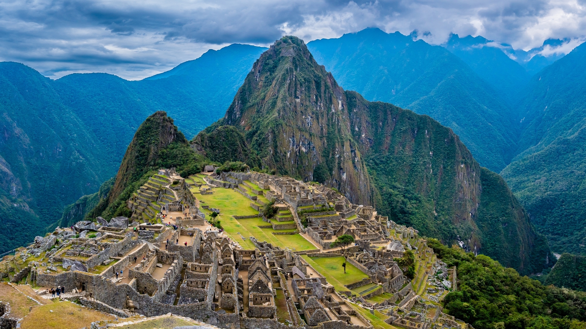 The image depicts Machu Picchu, an ancient Incan city located in the Andes mountains of Peru. It showcases the ruins of stone structures set against a backdrop of lush green mountains and a cloudy sky, highlighting the historical significance and breathtaking scenery of this UNESCO World Heritage site.