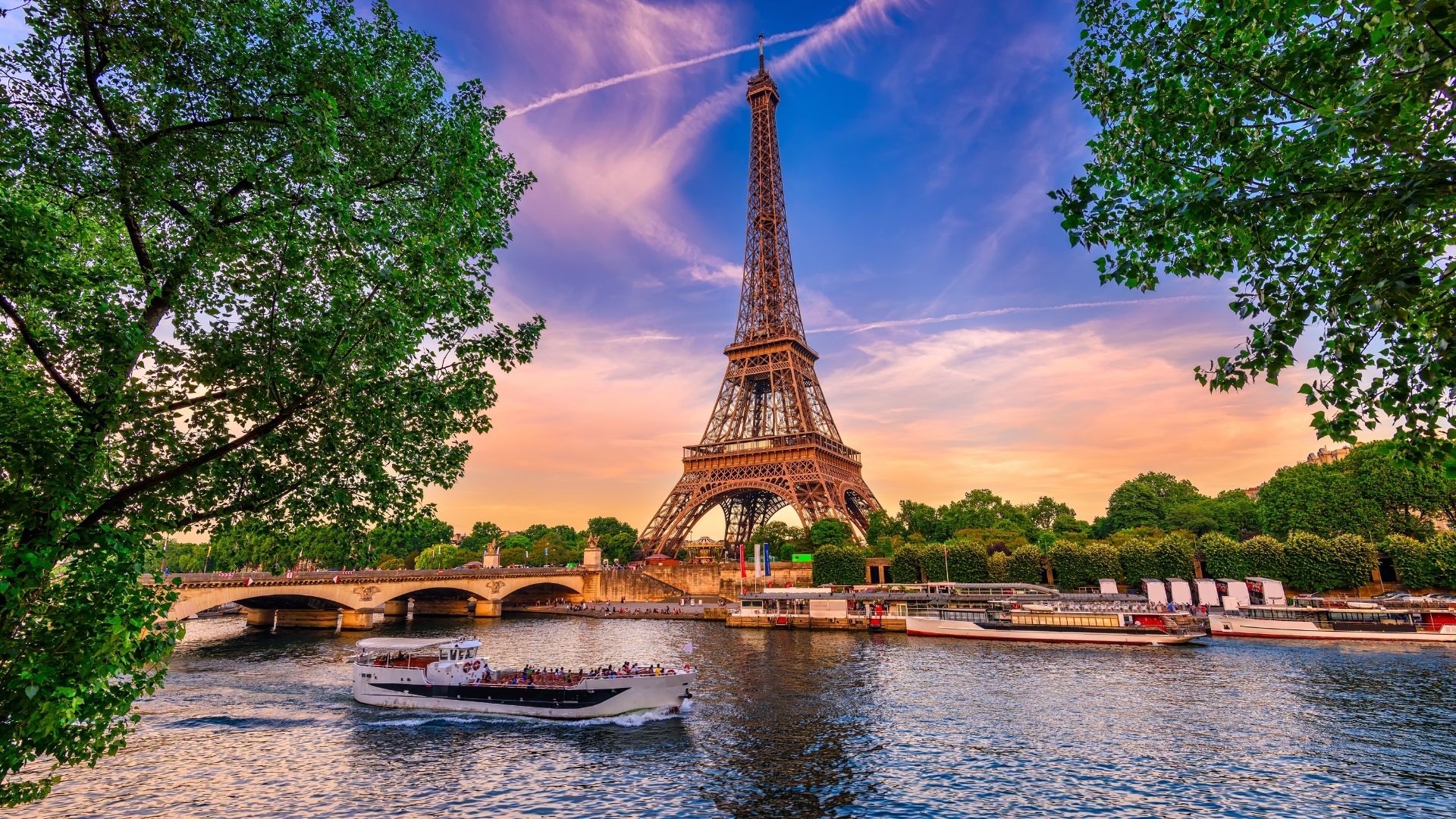 The image depicts the Eiffel Tower in Paris, framed by lush green trees, with the Seine River flowing in the foreground. A boat is seen cruising on the river, and the sky is painted with warm sunset colors, creating a picturesque scene of this iconic landmark.