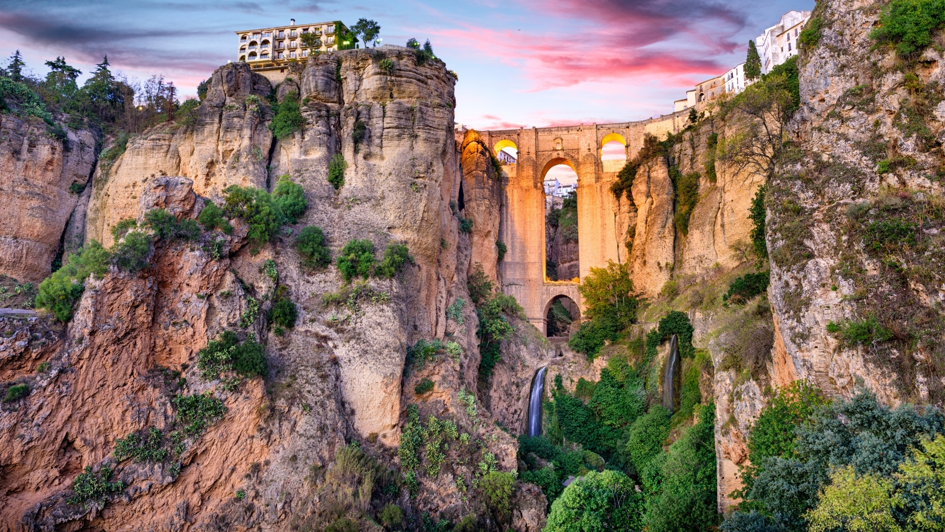 The image depicts the Puente Nuevo Bridge in Ronda, Spain, which is a stunning stone bridge spanning a deep gorge. The bridge features multiple arches and is surrounded by rocky cliffs and lush greenery, with a picturesque sky at sunset in the background.