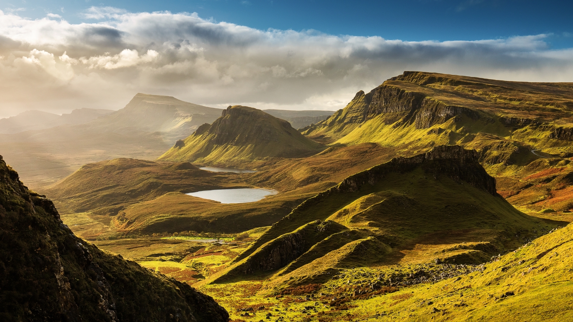 A scenic view of the Quiraing mountains in the Isle of Skye, showcasing lush green hills, a tranquil lake, and dramatic cloud formations in the sky.