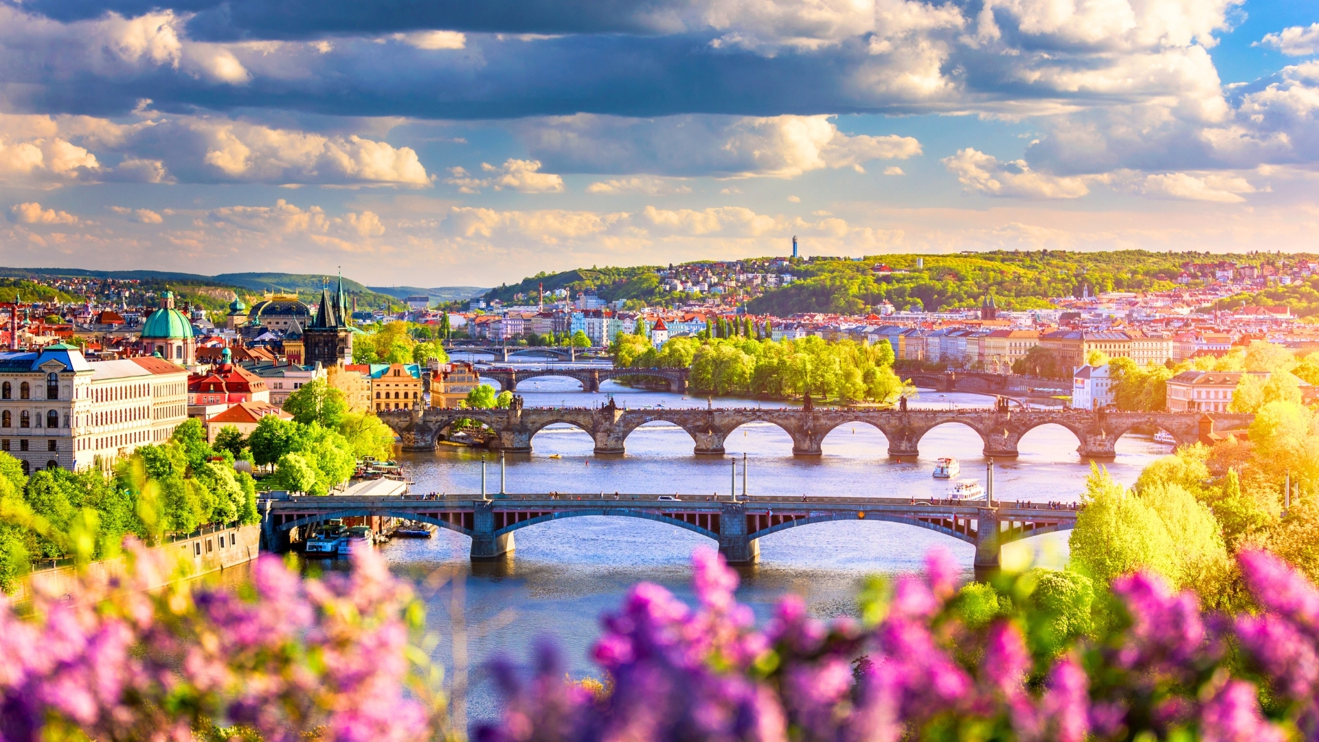 The image showcases a scenic view of Prague, featuring the Vltava River with multiple bridges, including the iconic Charles Bridge. The city is adorned with lush greenery and colorful buildings under a partly cloudy sky, creating a vibrant atmosphere.