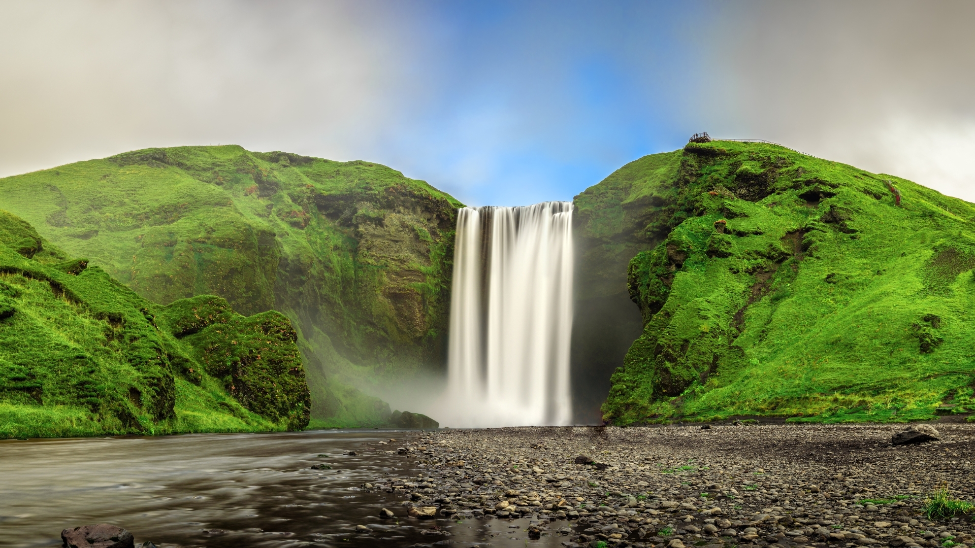 The image depicts the Skogafoss waterfall in southern Iceland, showcasing a powerful cascade of water falling from a height, surrounded by lush green hills and a clear blue sky. The scene captures the natural beauty and grandeur of the waterfall, with a river flowing in the foreground and rocky terrain.