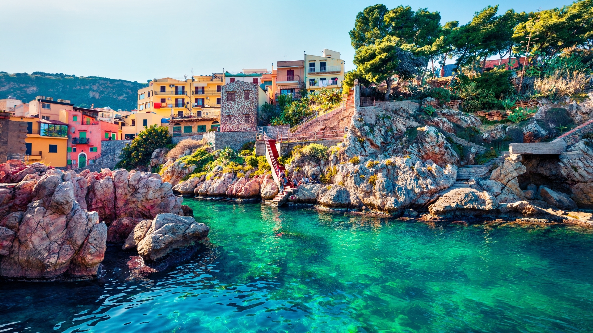The image depicts a picturesque view of Sant Elia village in Sicily, showcasing colorful buildings along a rocky coastline with clear, azure waters. The scene captures the beauty of nature and architecture, highlighting the vibrant colors of the houses against the backdrop of lush greenery and rocky formations.