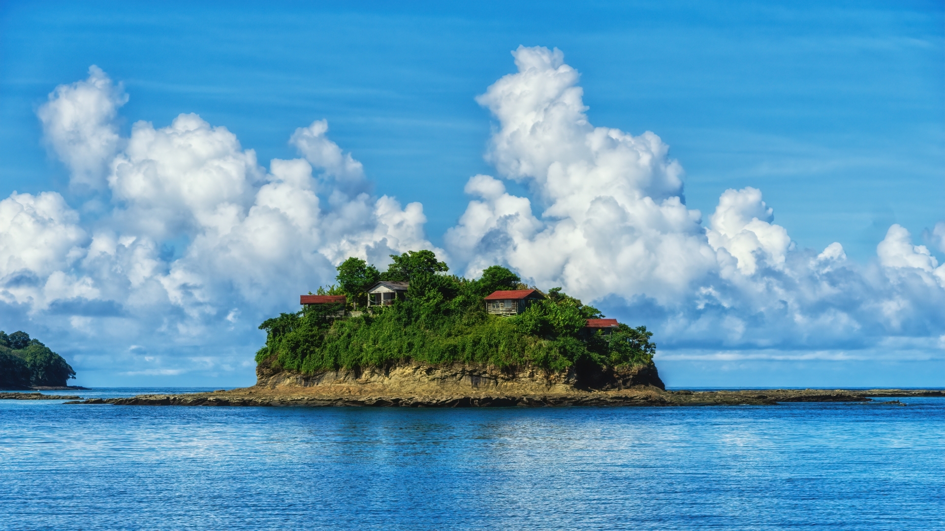 The image depicts a small, lush green island with several houses on it, surrounded by calm blue waters and a bright blue sky filled with fluffy white clouds. This serene scene captures the beauty of a tropical paradise, likely part of the Pearl Islands archipelago in Panama.