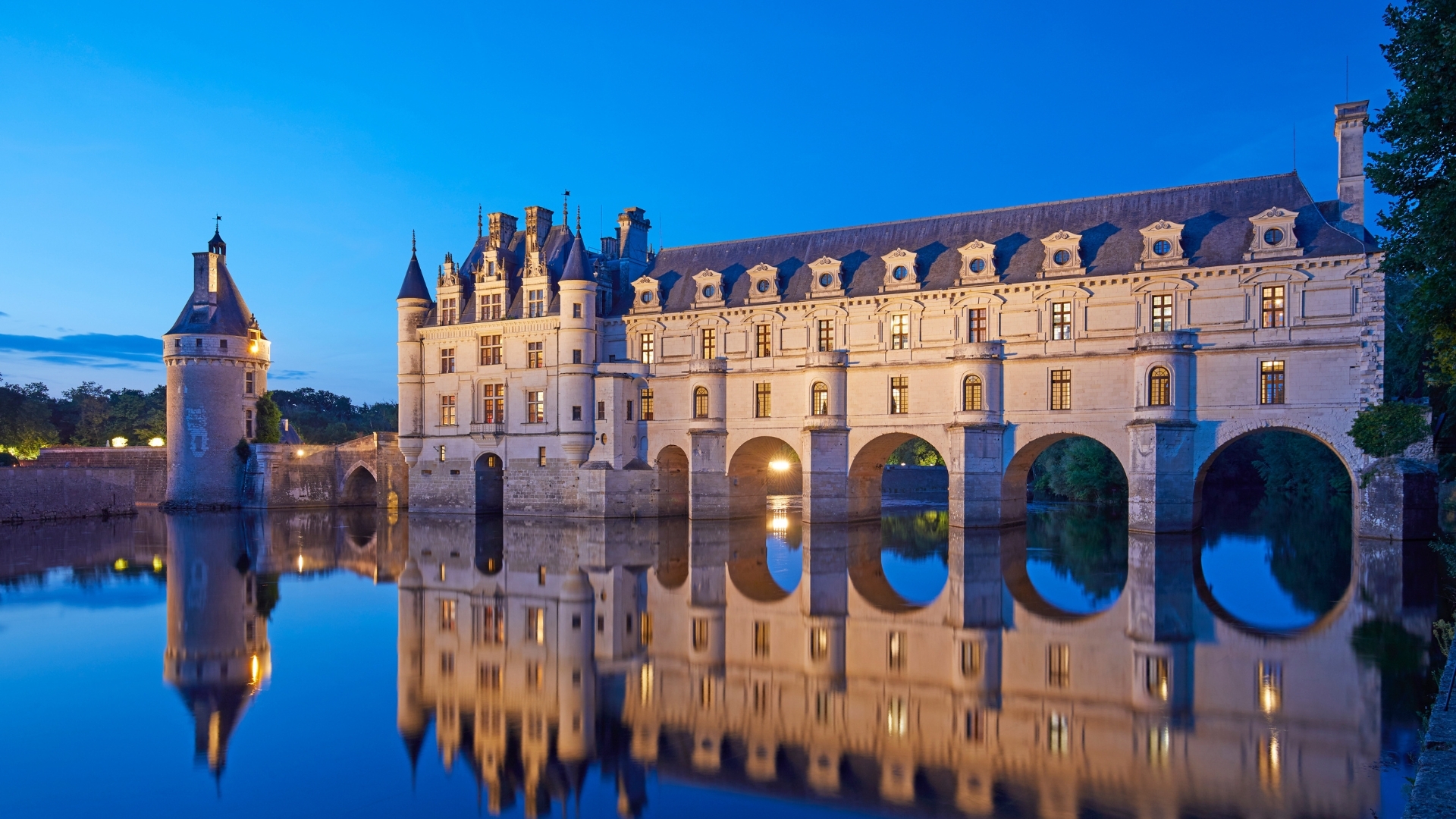 The image depicts the historical Chateau de Chenonceau, located on the Cher river in the Loire Valley, France. The castle is beautifully illuminated at dusk, reflecting in the calm waters below, showcasing its elegant architecture and surrounding nature.