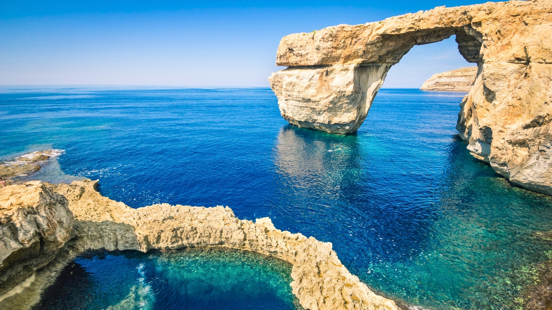 The image depicts the Azure Window, a natural limestone arch located on the island of Gozo in Malta, surrounded by clear blue waters and rocky cliffs.