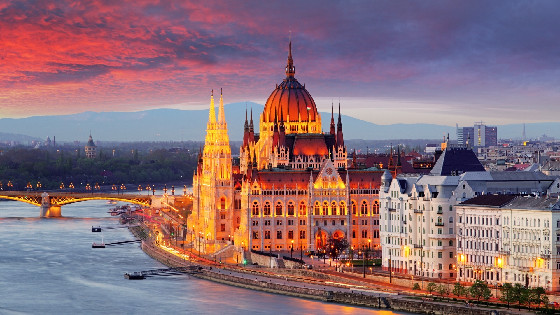 The image depicts the Hungarian Parliament building in Budapest at sunset, showcasing its stunning architecture illuminated by warm lights against a vibrant sky filled with shades of orange and purple.