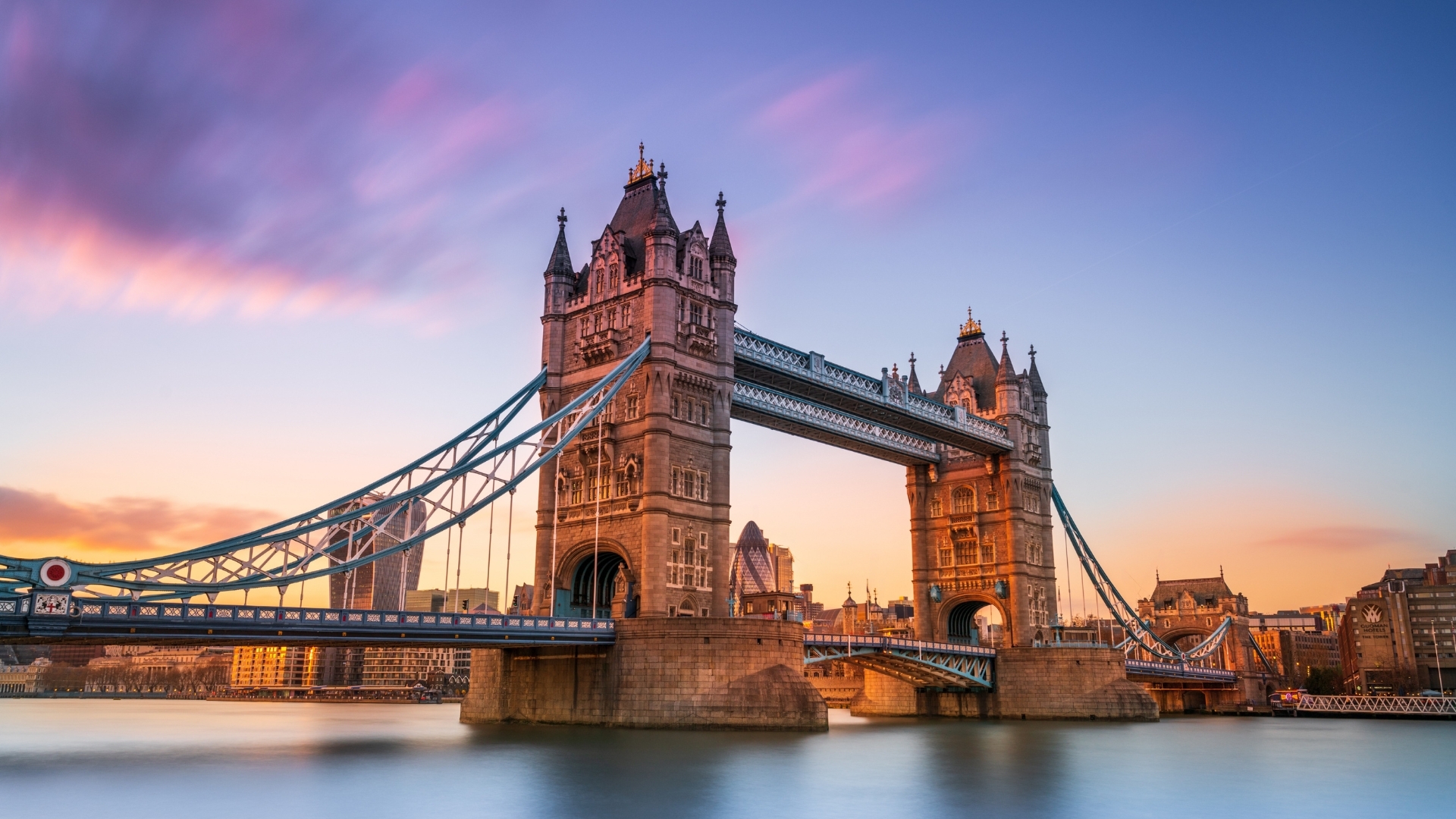 The image depicts the iconic Tower Bridge in London at sunset, showcasing its majestic towers and intricate design against a colorful sky. The bridge is illuminated by the warm hues of the setting sun, reflecting on the water below, with modern buildings in the background.